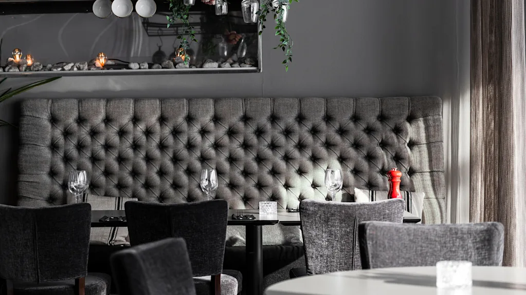Gray upholstered bench with tufted design; paired with dining tables set with glasses and a red pepper mill. Hanging lamps and greenery provide a modern atmosphere. 