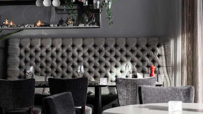 Grå polstret benk med tuftet design; sammen med spisebord dekket med glass og en rød pepperkvern. Hengelamper og grønt skaper en moderne atmosfære. 