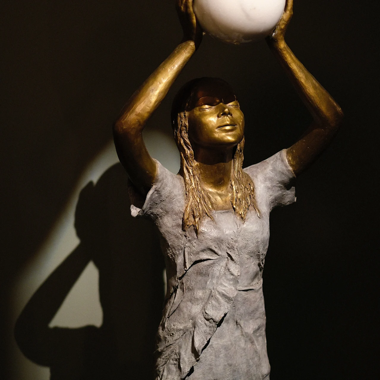 A statue of a figure is holding a white sphere overhead. The figure is in a grey textured outfit, and stands against a dark background.