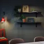 A wall with two shelves holds plants and framed art above a table. A red chair sits below, beside a red lamp, in a softly lit room.