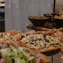 Pasta sallad med oliver ligger i en pappersklädd skål på en buffé. I närheten finns skålar med hackade grönsaker och blandade sallader, som förstärker en mysig matmiljö.