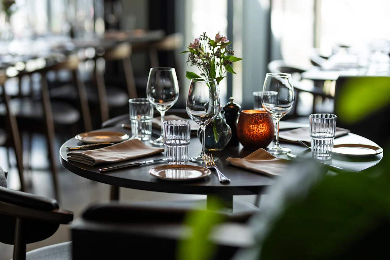 Et vakkert dekket restaurantbord med glass, bestikk, servietter, blomster og et glødende lys, klart for middag.