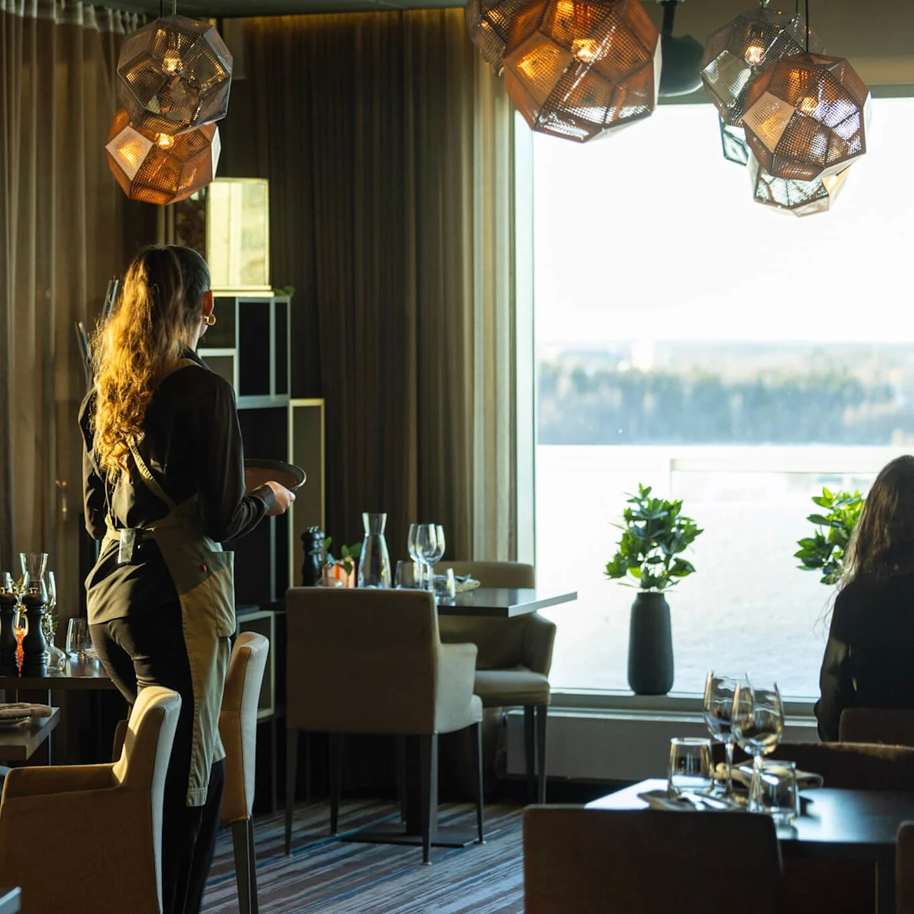 En servitör bär tallrikar och går genom en sofistikerad restaurang med elegant dukade bord. Restaurangen har geometriska hänglampor och stora fönster med naturskön utsikt.