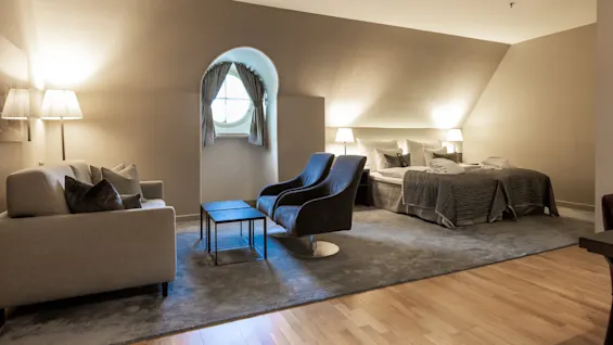 A spacious hotel room features a seating area with a sofa and chairs, two beds with bedside lamps, and a round window, creating a cozy, softly lit atmosphere.