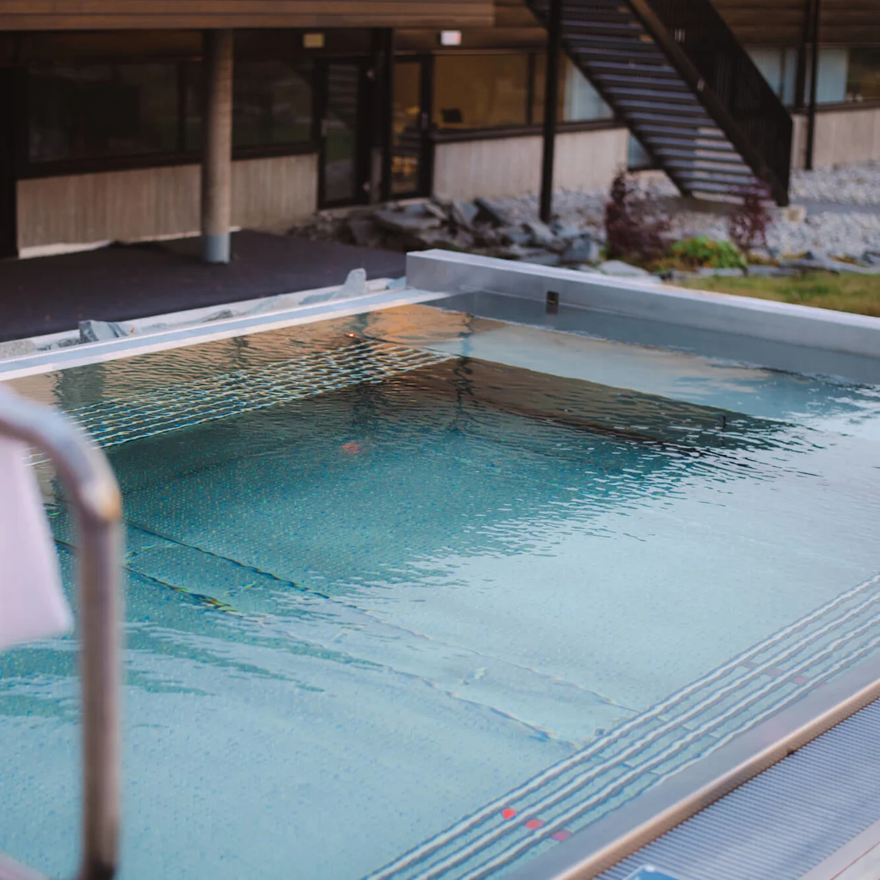 En pool reflekterar solljuset, omgiven av en byggnad med lägenheter. En vit handduk hänger över ett metallräcke vid poolen.