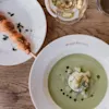 Grøn suppe pyntet med fløde og krydderurter i en tallerken mærket "WISBY HOTELL," ved siden af et spyd på en lille tallerken, altsammen på et træbord med et glas hvidvin.
