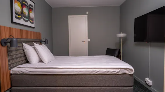 En pent oppredd seng står i sentrum av et minimalistisk soverom med grå vegger, flankert av en vegglampe. En moderne gulvlampe står ved siden av en stol og en veggmontert TV.