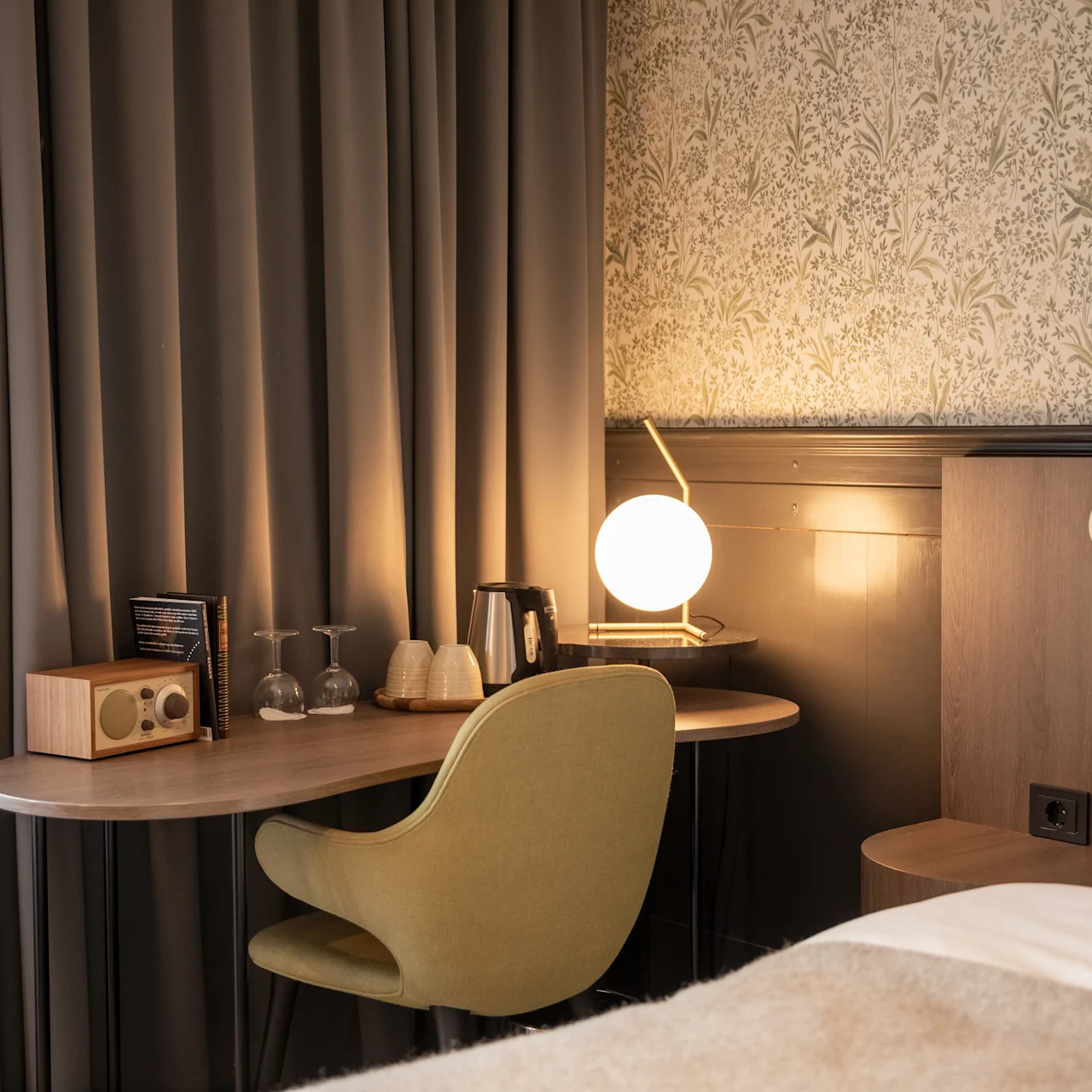 Ett skrivbord med en grön stol har en radio, glas och en sfärisk lampa, bredvid en säng. Mörka gardiner och ett mönstrat tapet skapar en mysig rumstämning.
