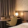 Et skrivebord med en grønn stol har en radio, glass og en sfærisk lampe, ved siden av en seng. Mørke gardiner og mønstret tapet skaper en koselig romatmosfære.