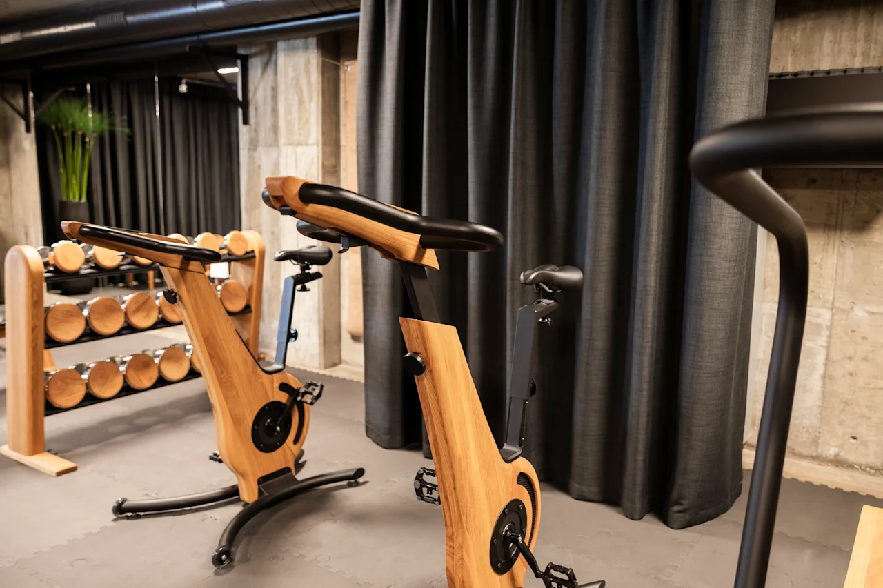 Träningscyklar med ramar av trä står still i ett gym, omgivna av svarta gardiner och ett ställ med hantlar.