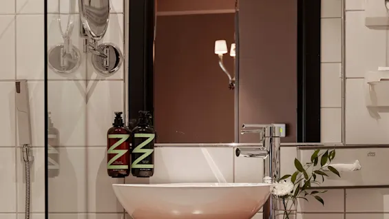 A round basin sits on a wooden shelf holding a white towel, with two bottles labeled "Z" beside a mirrored wall in a tiled bathroom; a vase with flowers is beside the tap.