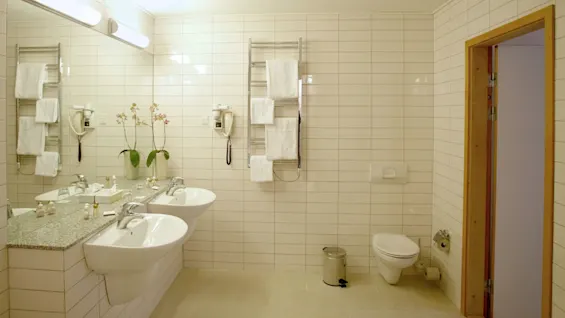 Double sinks rest on a granite countertop, holding toiletries and orchids, in a bright, tiled bathroom featuring towel racks, a toilet, and a small trash bin near an open wooden door.
