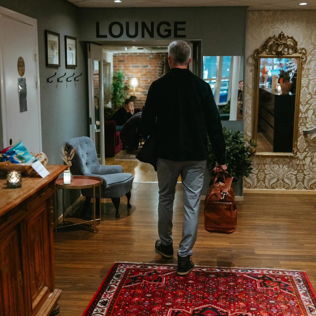Mies kävelee kohti 'LOUNGE' -merkittyä huonetta, kantaen laukkua, tyylikkäästi sisustetussa käytävässä, jossa huonekaluja ja peili.