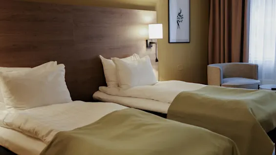Two neatly made twin beds rest against a wooden headboard, covered with green blankets. A wall lamp illuminates an adjacent chair and curtained window, creating a cozy hotel room atmosphere.