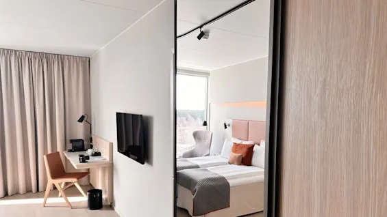 A modern hotel room features a neatly arranged bed beside a window. A desk with a chair and a wall-mounted TV are against the adjacent wall. Light wood flooring and soft lighting enhance the minimalist design.