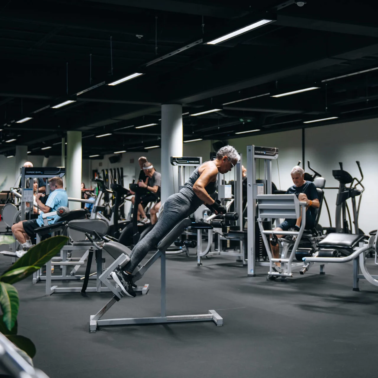 En person tränar på en ryggträningsmaskin, omgiven av andra gymbesökare som använder olika utrustningar. Gymmet har en modern inredning med synliga träningsmaskiner och en digital klocka som visar "13:39."
