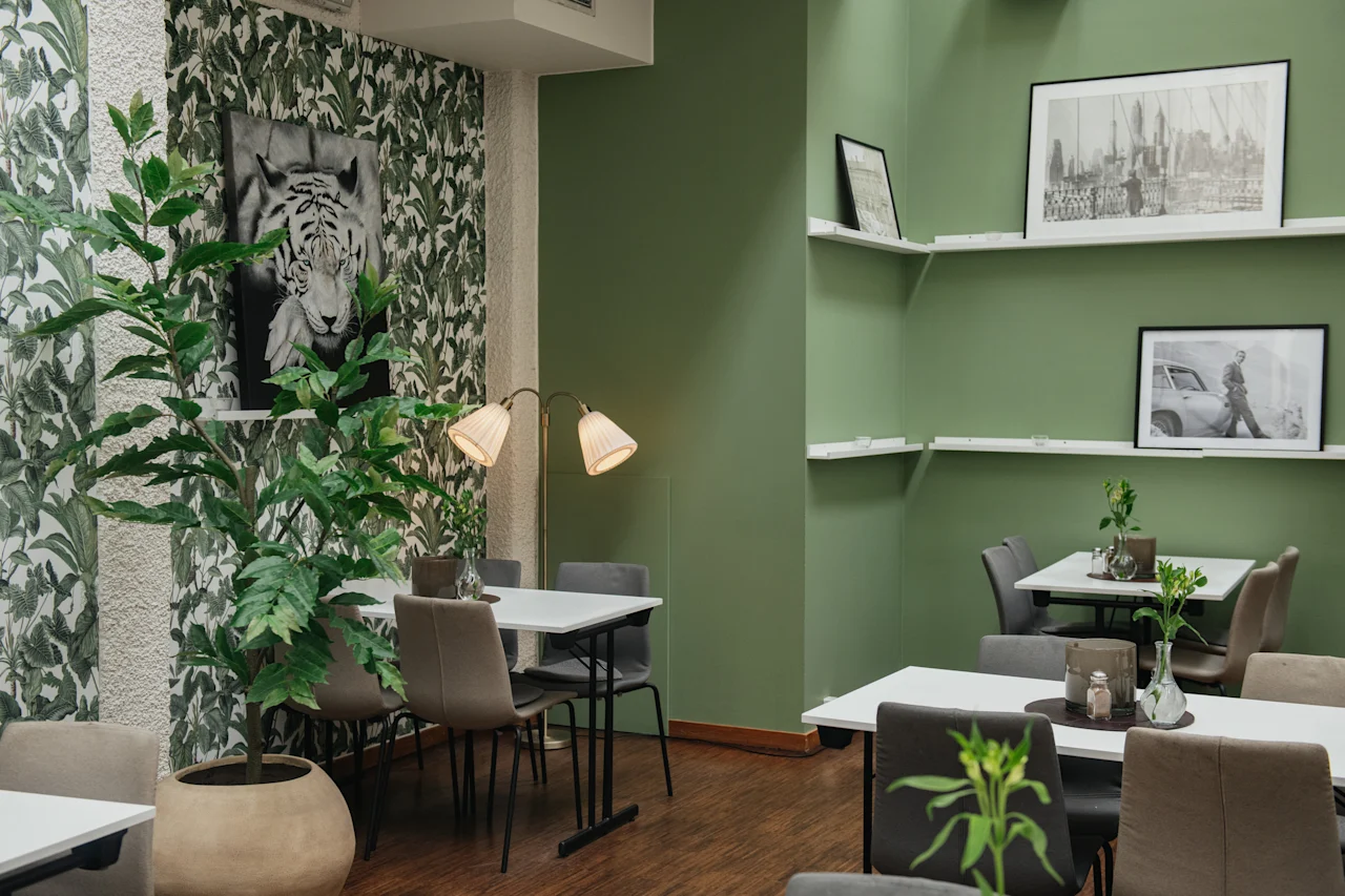 Bord och stolar är arrangerade i ett grönt väggkafé prytt med lövverkstapeter och monokrom konst. En krukväxt och mjuk belysning förstärker den välkomnande atmosfären.
