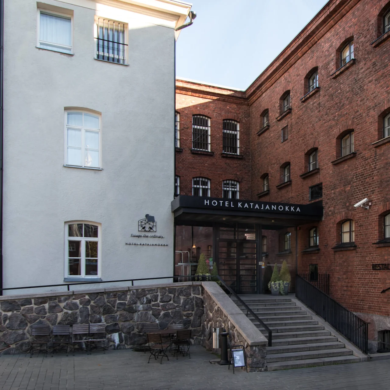 Brick hotel building with entrance canopy, stone stairs leading up, adjacent white building, serene courtyard setting. Text: "HOTEL KATAJANOKKA" and "Escape the ordinary".