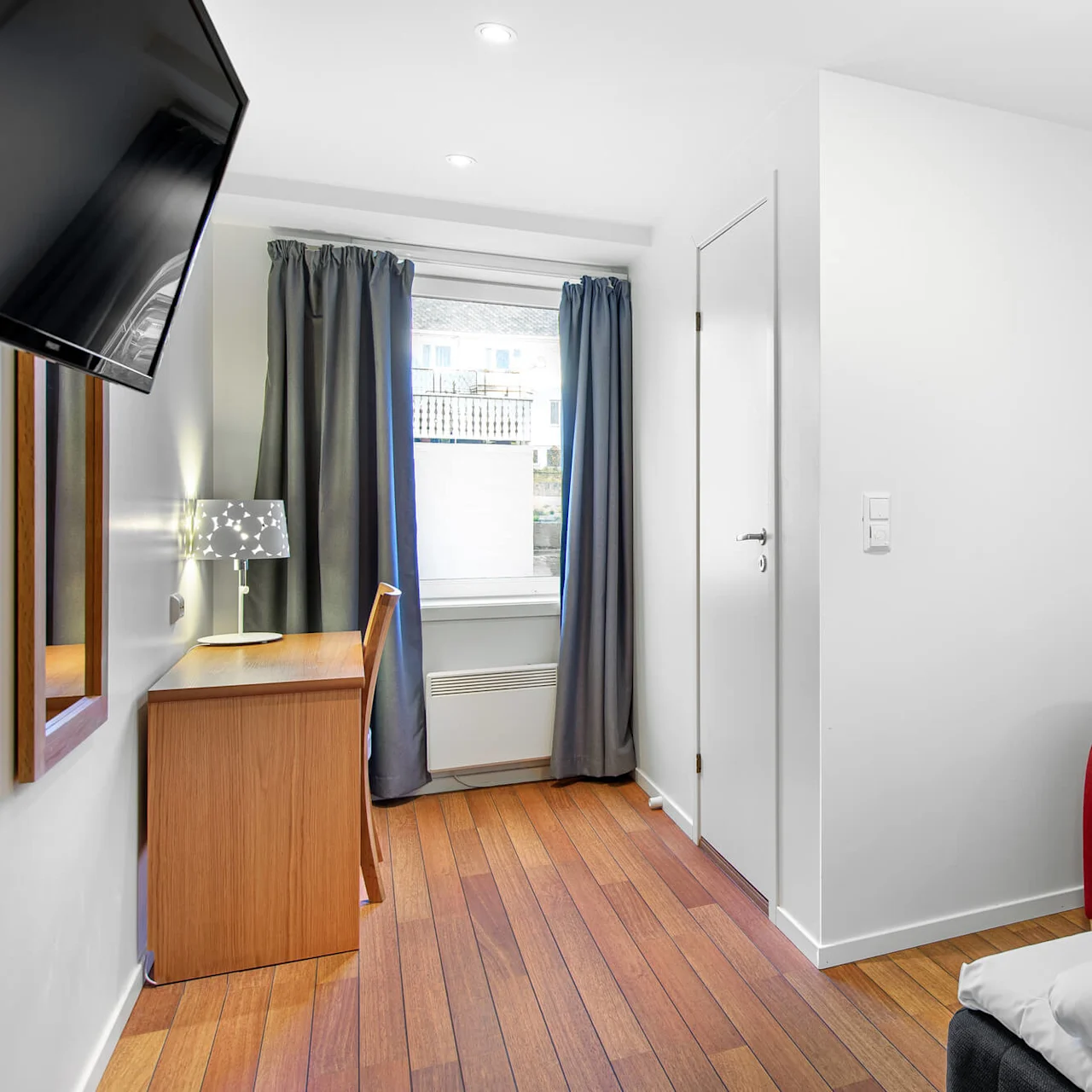 Rød lenestol ved siden av en seng i et hotellrom, supplert med et skrivebord under et vindu med gardiner og en mønstret vegg.