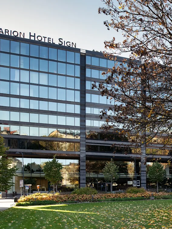 Modern hotellbyggnad i glas bredvid en gron park med trad pa en solig dag