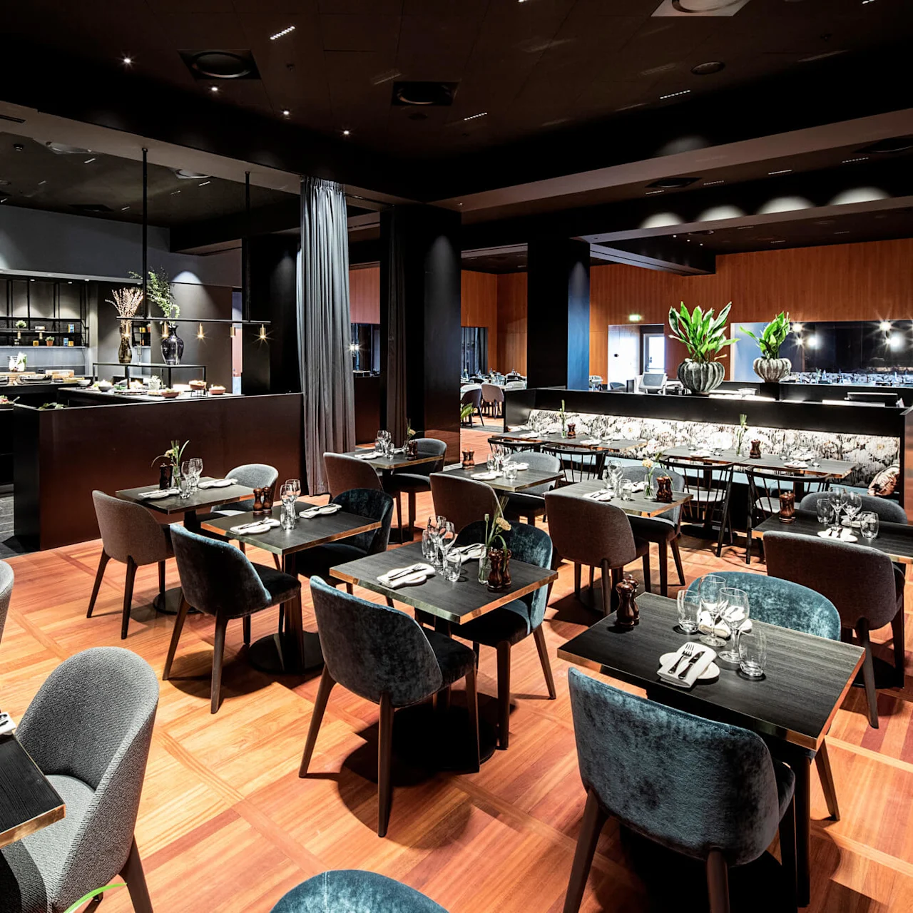 En rymlig restaurang med eleganta bord dukade med glas och servetter, omgiven av moderna stolar. I bakgrunden syns ett bufféområde med dekorativa växter och maträtter.