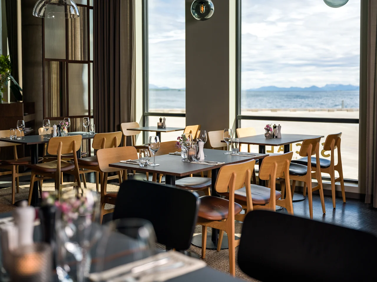 En lys restaurant på Quality Hotel Harstad med bord dekket for bespisning, med utsikt over vann og fjell.