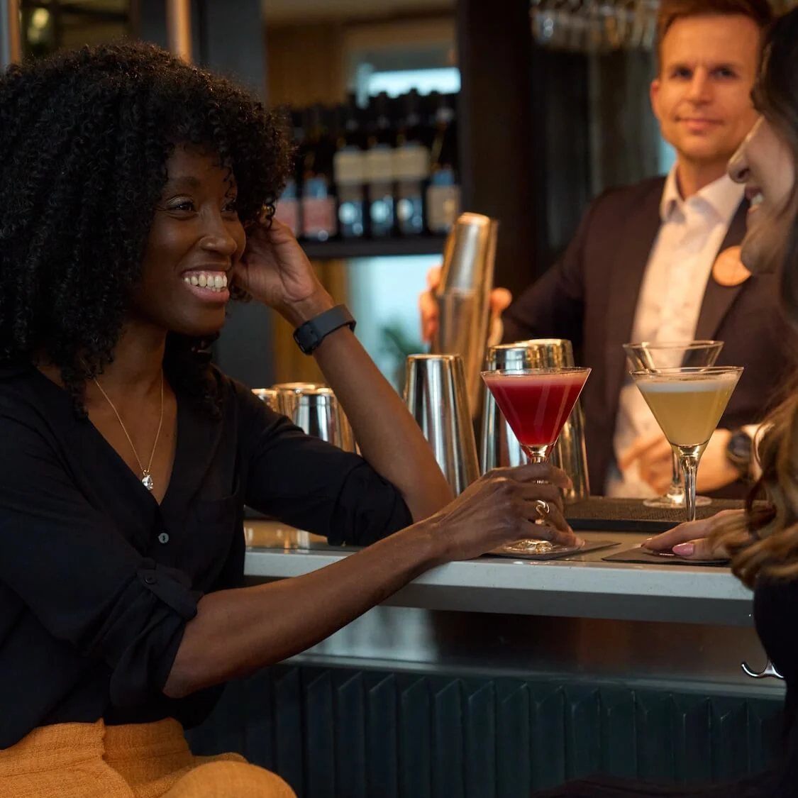 A woman smiles while holding a red cocktail glass, seated at a stylish bar counter beside a friend, with a bartender preparing drinks in the background.