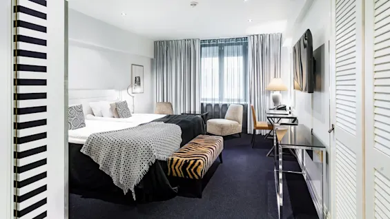 Et hotellrom har en pent oppredd seng med mønstret sengetøy, en benk med zebramønster, og en sittegruppe med stoler og skrivebord. Gardiner dekker et stort vindu.