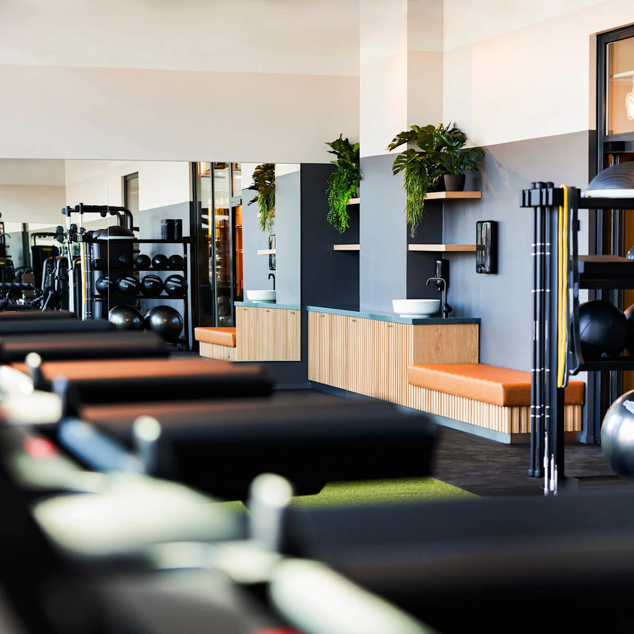 Løbebånd strækker sig på en række, vendt mod træningsudstyr og spejle i et elegant, moderne fitnesscenter med lyse trædetaljer og potteplanter.