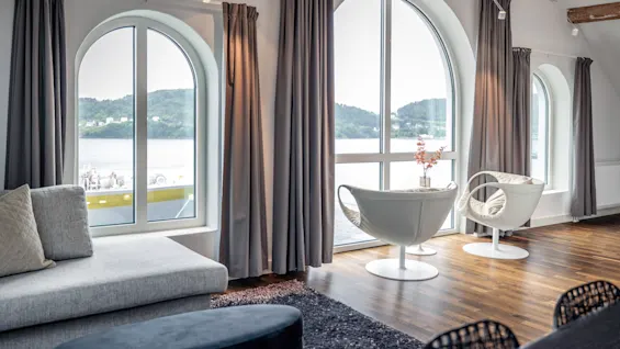 Två moderna vita stolar står vid stora välvda fönster med utsikt över vattnet. Grå gardiner hänger bredvid bekväma sittmöbler i ett elegant rum med trägolv.