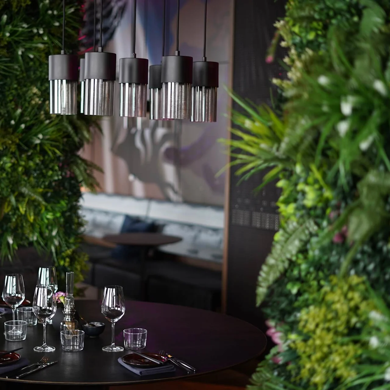 Moderne hengelamper henger over et rundt spisebord med glass og bestikk, omgitt av frodige grønne vegger i en koselig, eksklusiv restaurantinnredning.