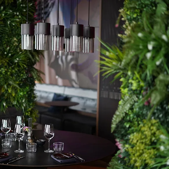 Moderna hänglampor hänger över ett runt matbord med glas och bestick, omgivna av frodiga gröna väggar i en mysig, exklusiv restaurangmiljö.