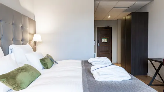 A neatly made bed with white linens and green pillows is against a gray headboard, beneath a wall lamp, in a hotel room with wooden door, closet, and table.