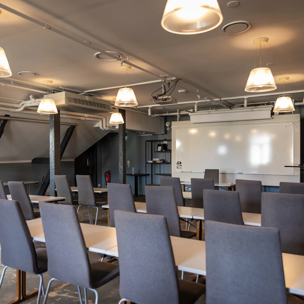 Klasseværelsesopsætning: Tomme grå stole vender mod et whiteboard i et loftsstillignende rum, oplyst af loftlamper. Borde er arrangeret til præsentationer eller foredrag.