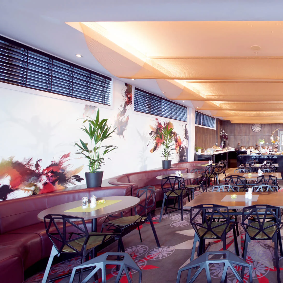 A modern restaurant with empty tables and chairs, large windows, and abstract wall art providing a bright and spacious dining atmosphere.