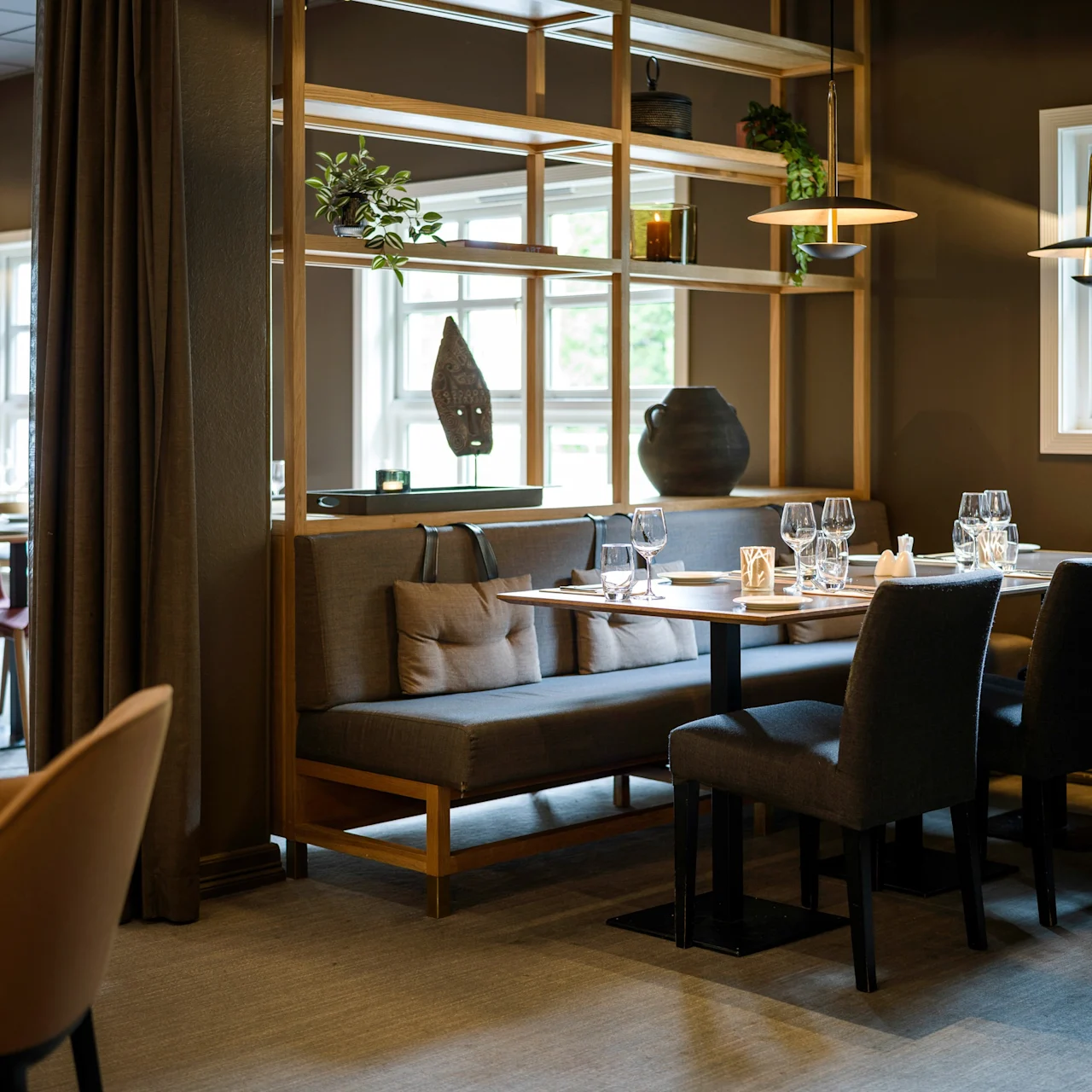 Elegant dining setup displayed on a wooden table surrounded by cushioned chairs, within a warmly lit restaurant interior, featuring decorative shelves and large windows.