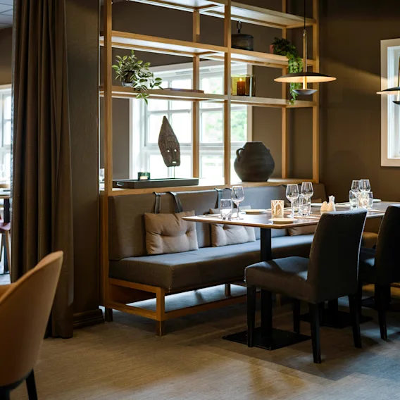 Elegant spiseoppsett vist på et trebord omgitt av polstrede stoler, innenfor en varmt opplyst restaurantinteriør, med dekorative hyller og store vinduer.
