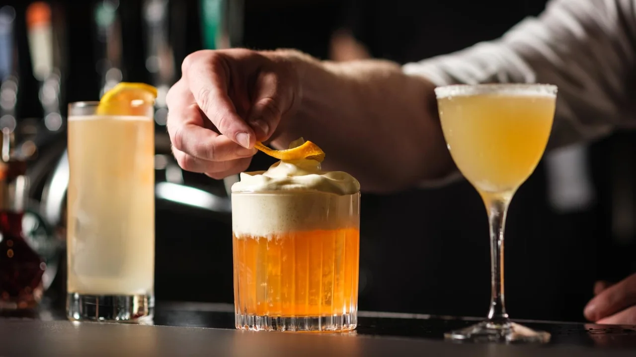 En bartender garnerar en skummig cocktail bland andra drycker på en mörk bardisk.