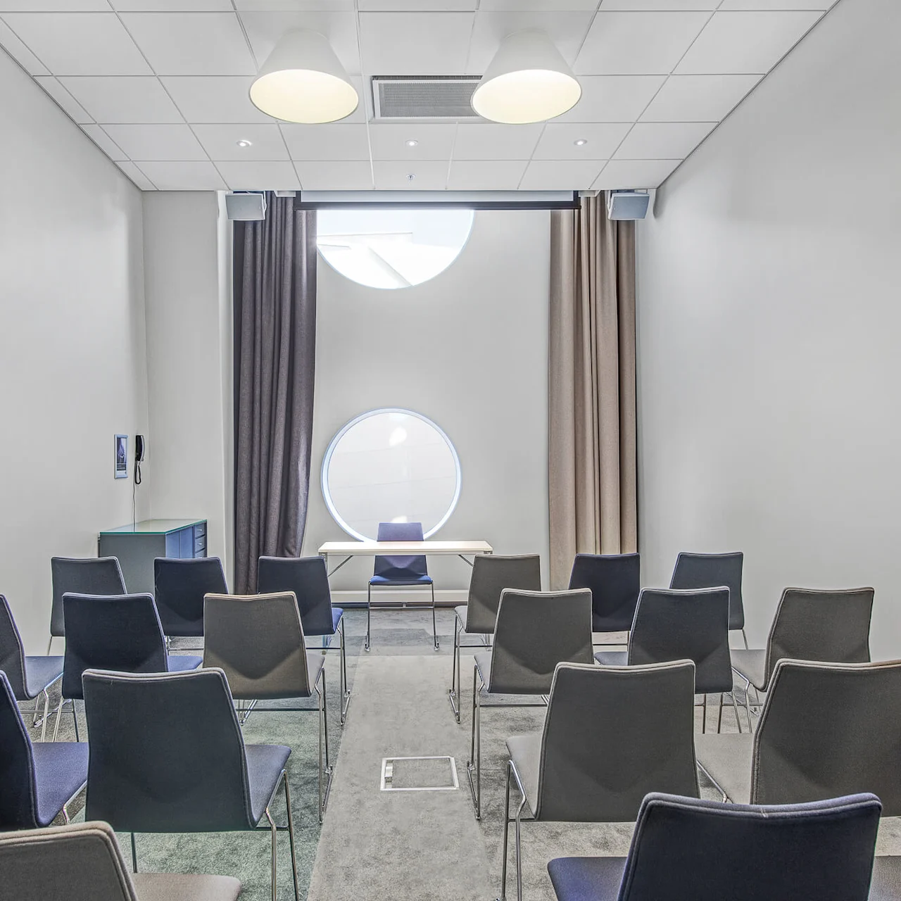 Rækker af stole vender mod et bord under runde vinduer, med gardiner og lys i loftet, i et lille, moderne konferencerum.