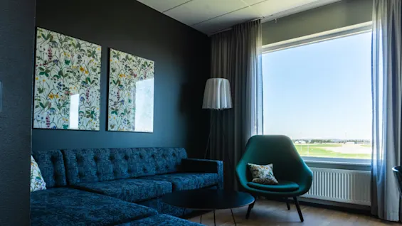 En mörkblå soffa står i ett vardagsrum med blommig konst på väggen, bredvid ett stort fönster med utsikt över ett soligt utomhusområde.