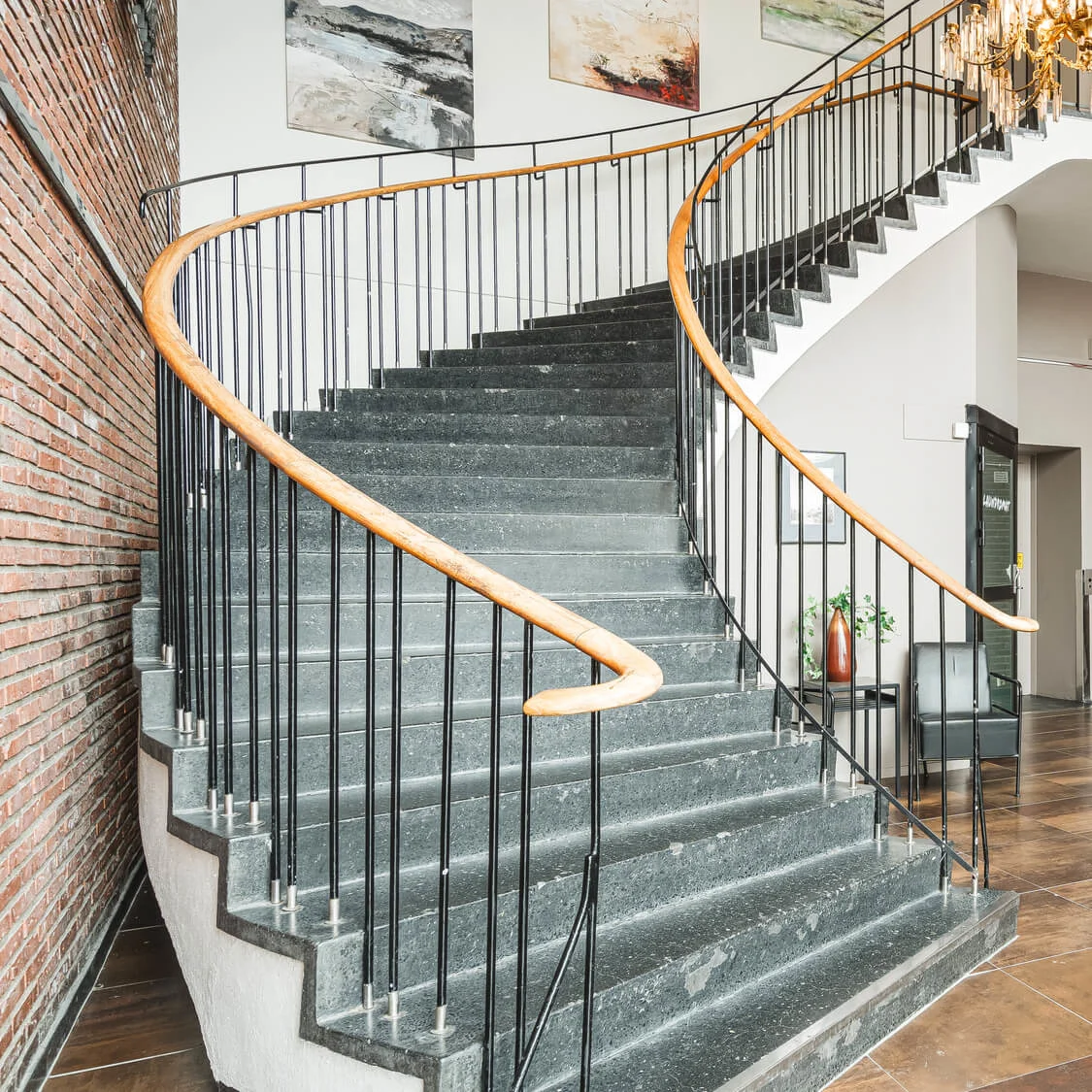 En böjd trappa med ett trähandtag stiger bredvid en tegelvägg, under en ljuskrona i en öppen, modern lobby med hissar och abstrakt konstverk.