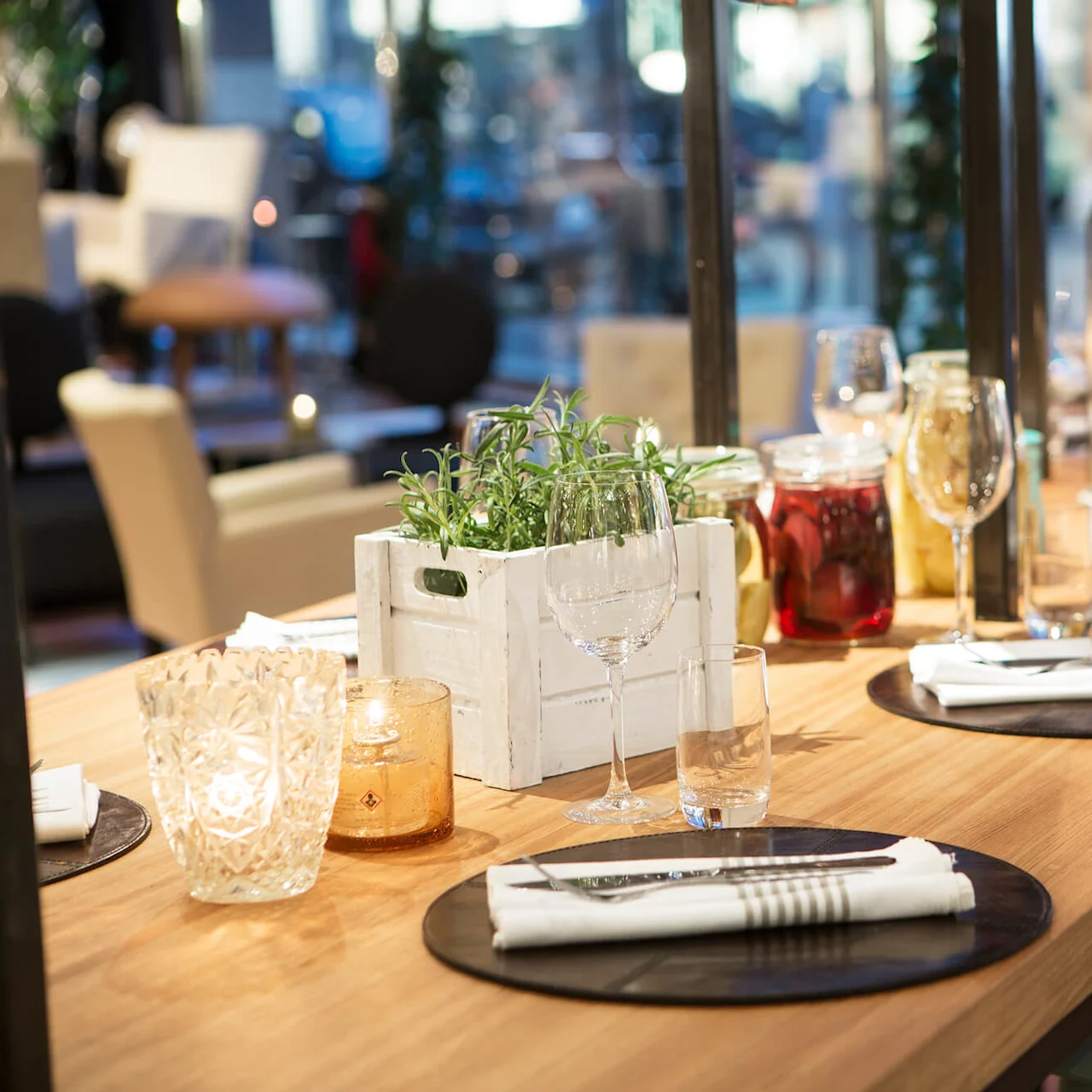 A wooden dining table is elegantly set with wine glasses, white napkins, and candles, surrounded by modern chairs in a softly lit restaurant environment.