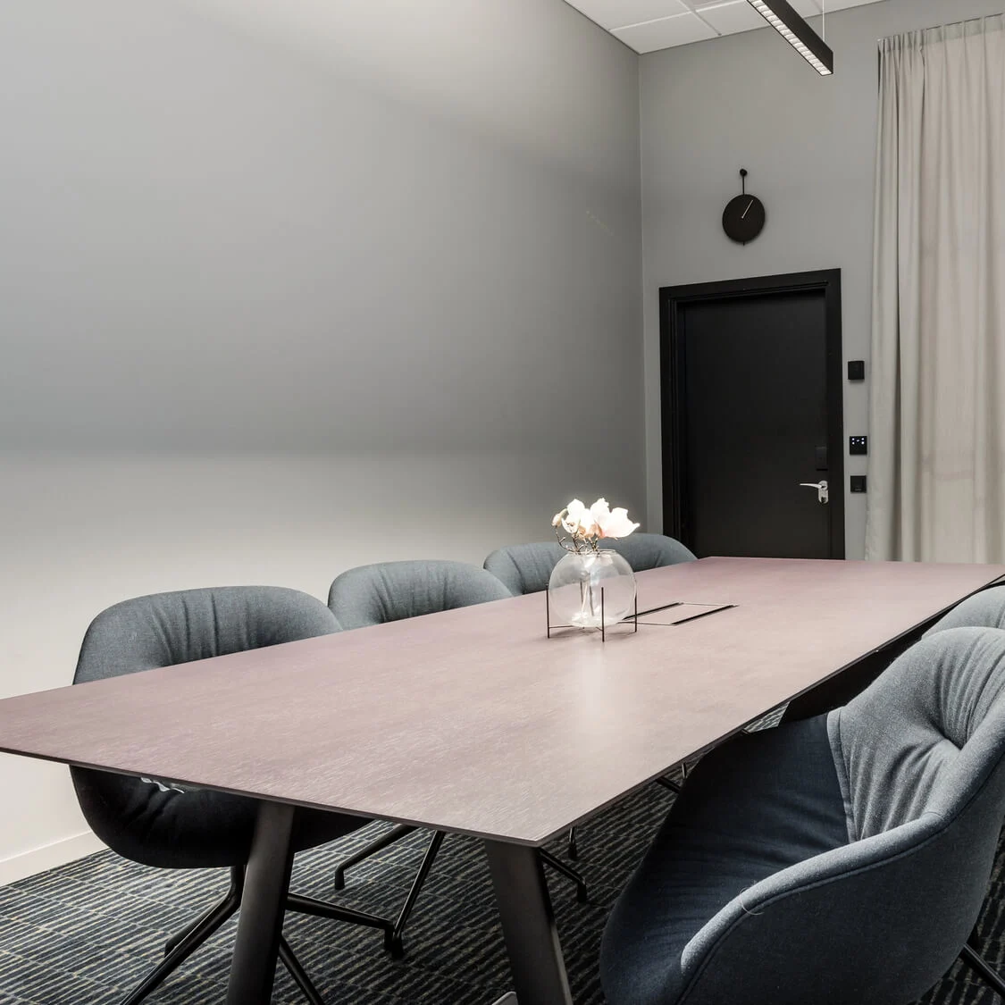 Et langt konferencetable omringet af seks polstrede stole, i et moderne mødelokale med grå vægge, en sort dør, et vægur og et mønstret tæppe.