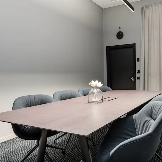 A long conference table surrounded by six upholstered chairs, in a modern meeting room with grey walls, a black door, a wall clock, and a patterned carpet.