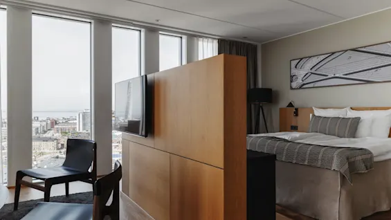 Et moderne hotellrom med stort vindu og byutsikt, med seng, sitteplasser og en trevegg med TV.