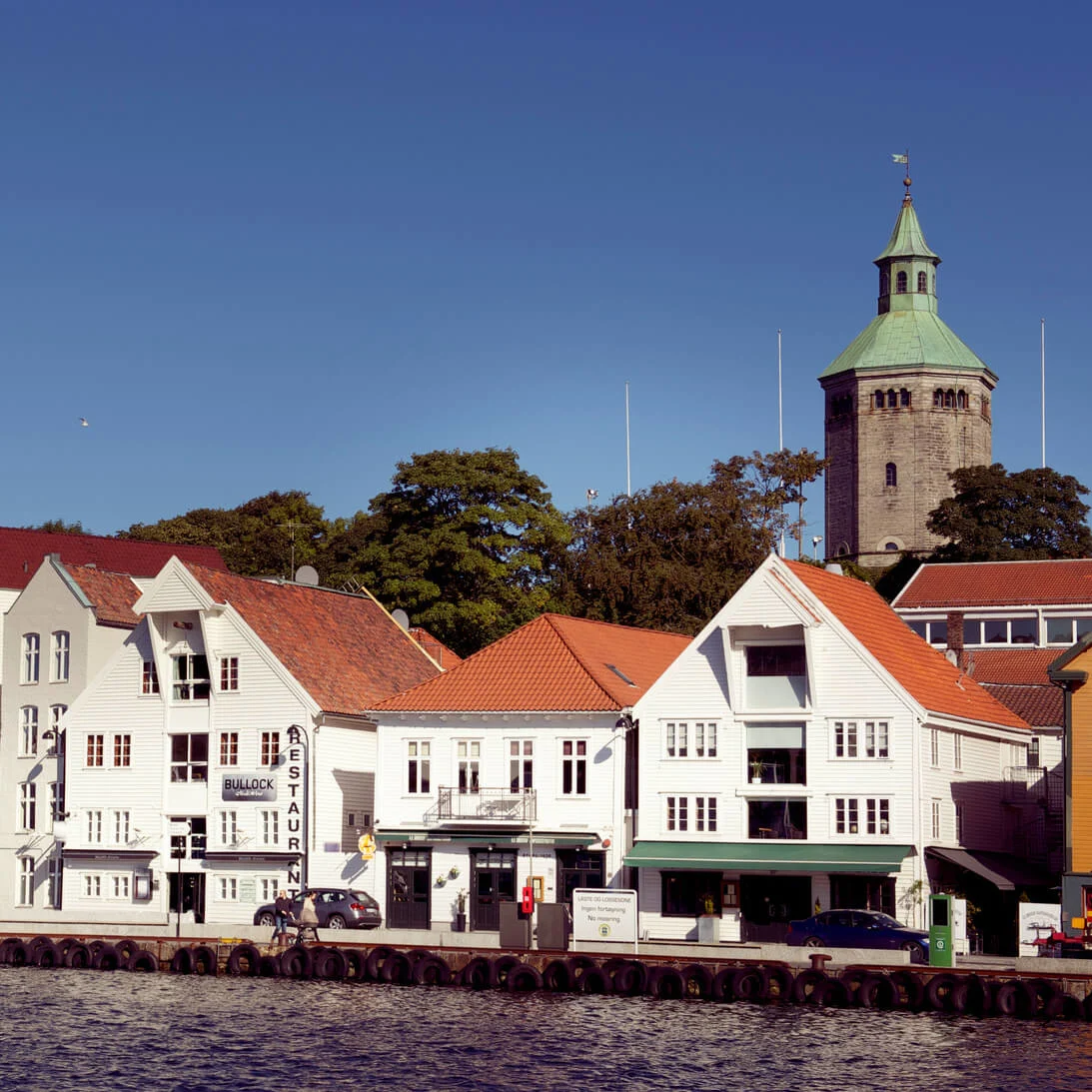 Traditional white wooden houses line the waterfront, under a clear blue sky with a green-topped tower in the background.