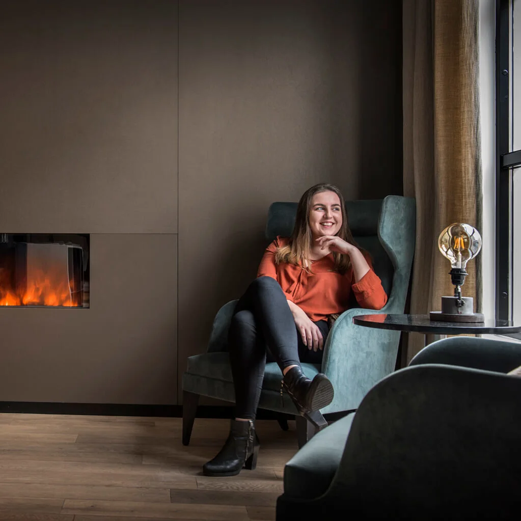 En kvinna sitter leende i en högryggad stol bredvid en väggmonterad eldstad i ett mysigt rum med bruna väggar och trägolv.