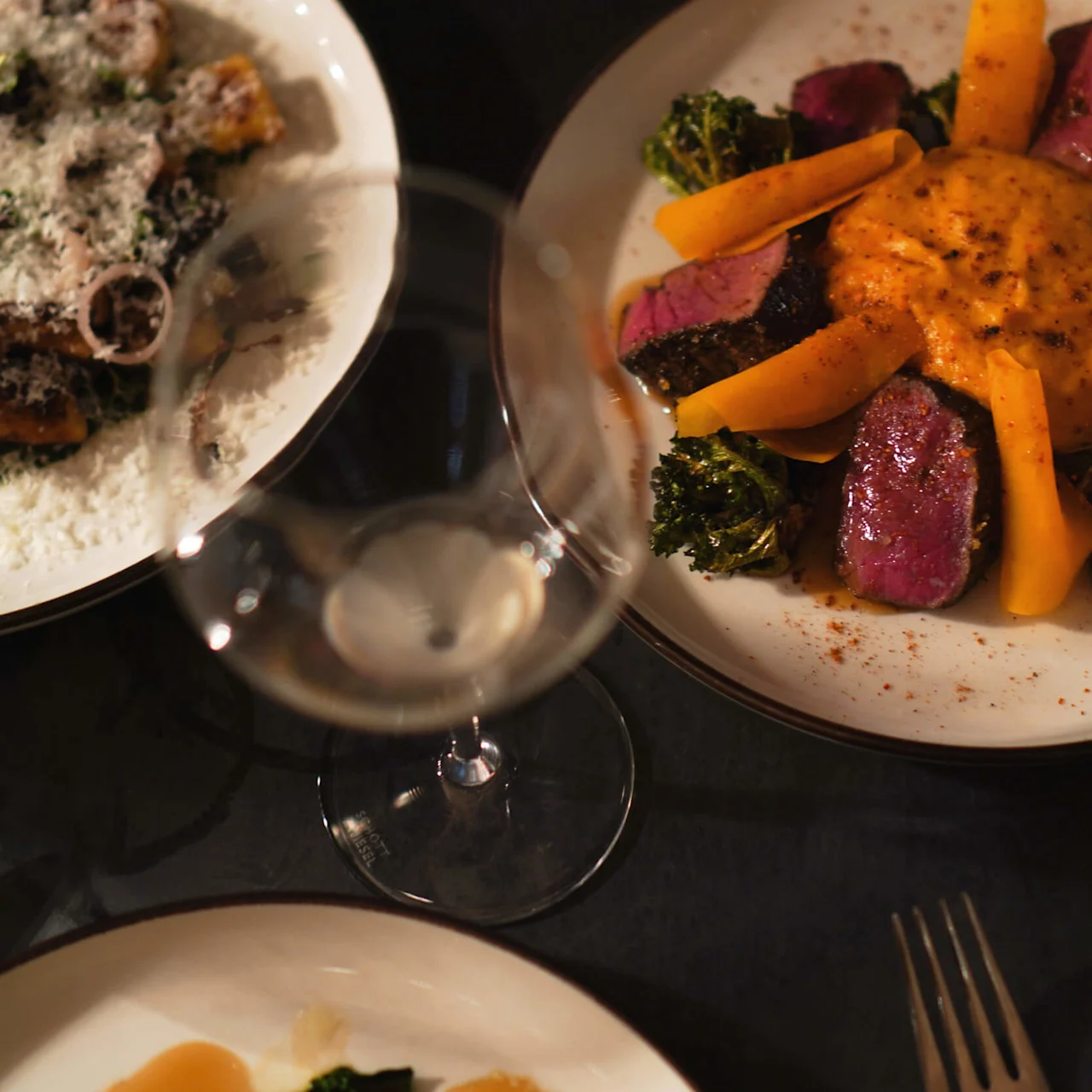 A plate holds sliced steak with bright yellow carrots and broccoli, centered around a creamy sauce. A wine glass is positioned above, and another plate is partially visible nearby.