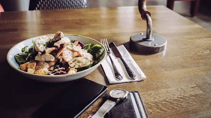 A salad bowl with chicken sits on a wooden table. A fork, knife, napkin, lamp, smartphone, wristwatch, and wallet are nearby. Background shows a dimly lit interior space.