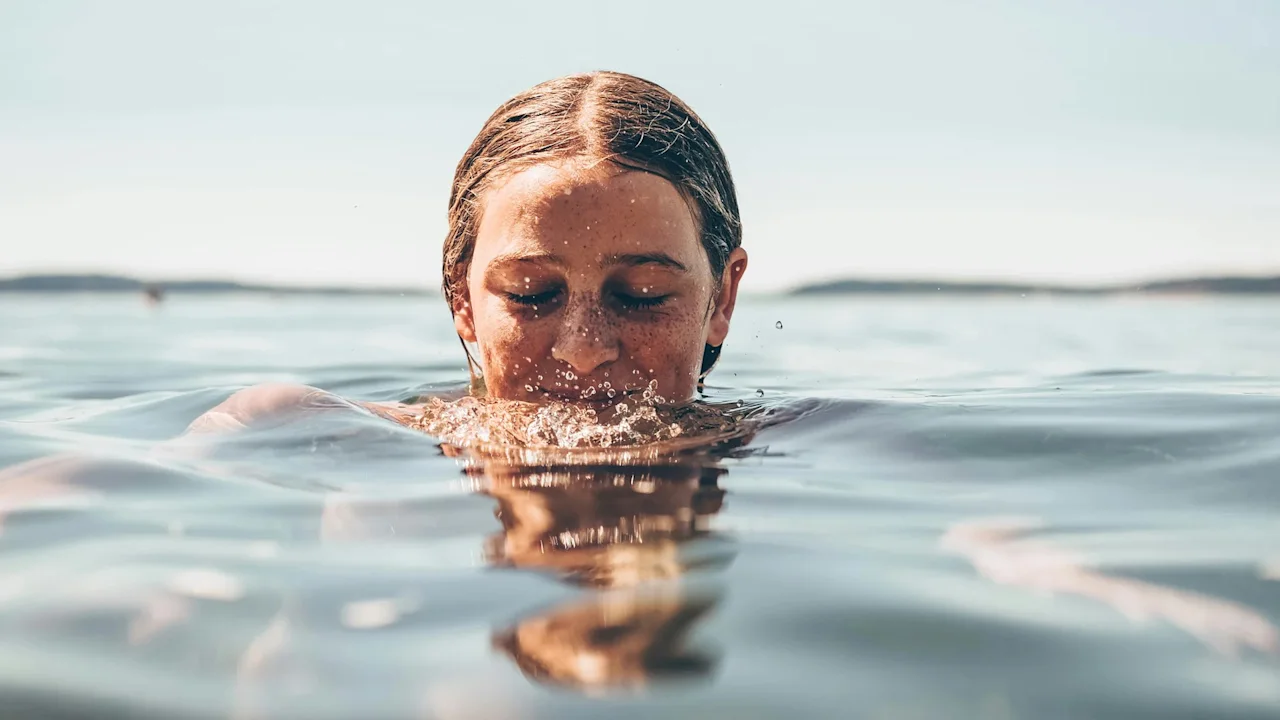 A person immersed in water exhales, creating small ripples, surrounded by a calm, expansive lake under a clear sky.